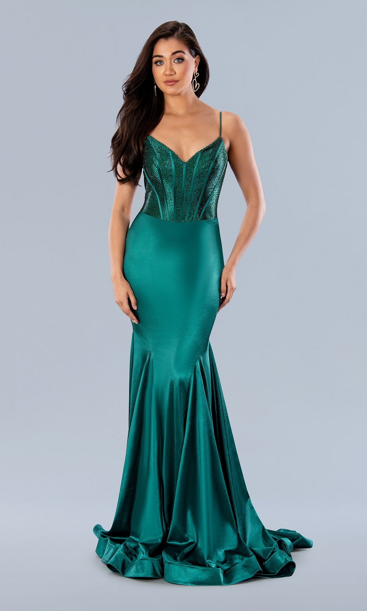 A woman wearing the Stella Couture 24198 Deep V-Back Long Prom Dress, featuring emerald green fabric, a beaded corset bodice, and thin straps, stands against a plain light gray background.